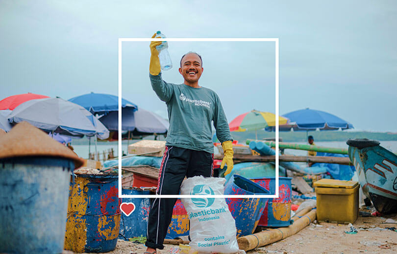 Plastic Bank male ocean hero ,smiling and holding a plastic bottle in front of Plastic Bank collection sac by the beach.